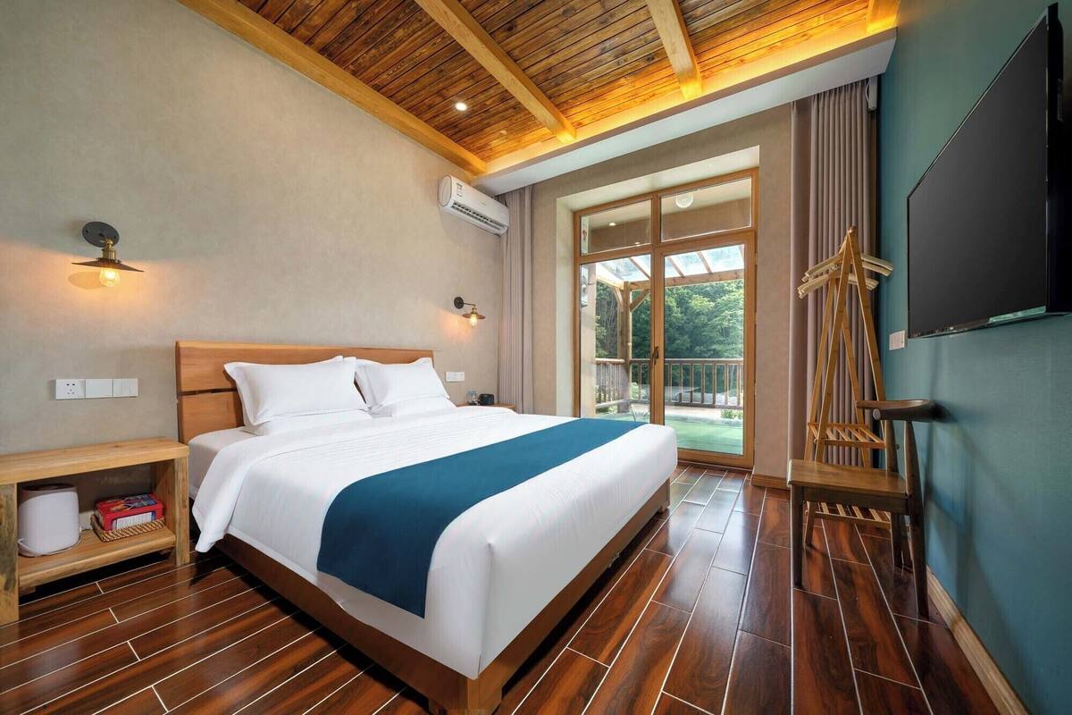 A hotel room with a wooden ceiling, a large bed with white bedding and a blue accent, a wooden nightstand, a TV, and a view of the outdoors through a glass door.