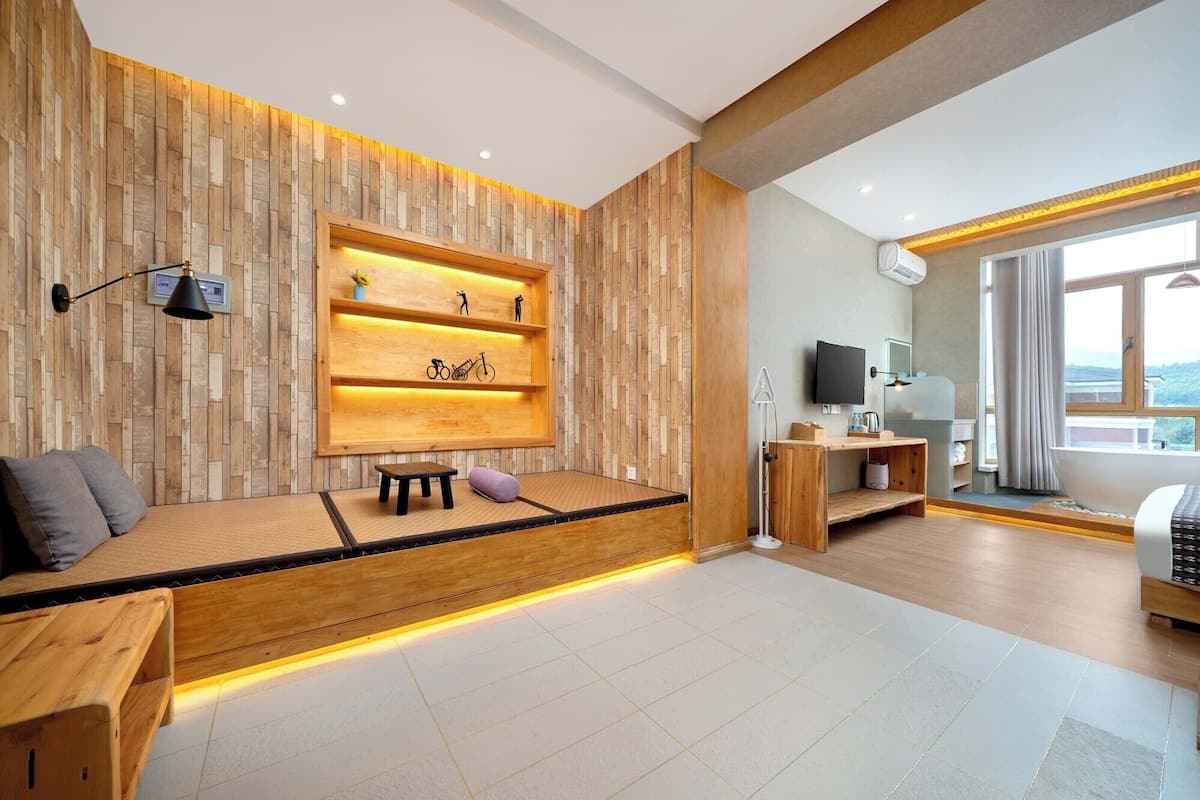 A modern hotel room with a wooden accent wall, a built-in bench with a shelf, a flat-screen TV, and a large window offering a view.