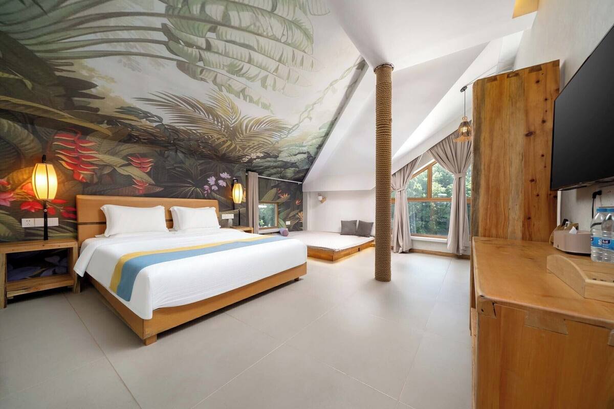 A spacious bedroom with a large bed, a ceiling mural, a wooden headboard, a nightstand, a bedside lamp, a TV, and a large window with curtains.