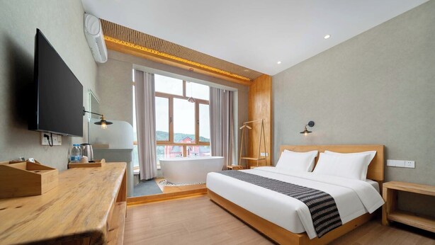 A modern hotel room with a large bed, a wooden desk, a flat-screen TV, a bathtub, and a large window offering a view of buildings. - Yabuli Liangqi Qingyun Homestay (Harbin)