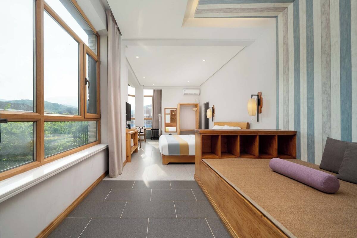 A modern hotel room with a large window, a bed, a seating area, and a bathroom.