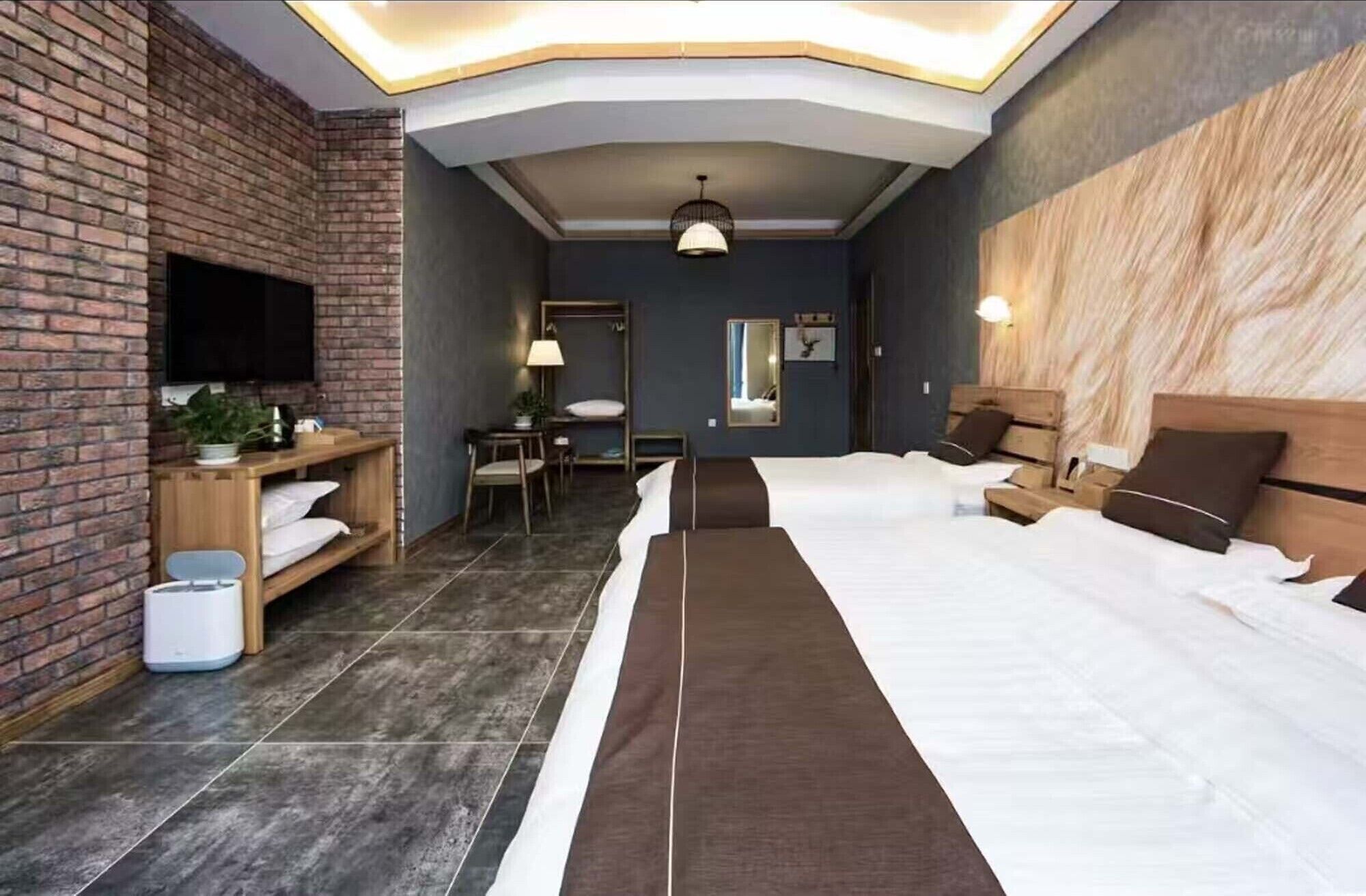 Una habitación de hotel moderna con una pared de ladrillo decorativa, un televisor de pantalla plana, un escritorio con silla, una cama con dos almohadas y una mesita de noche con lámpara.