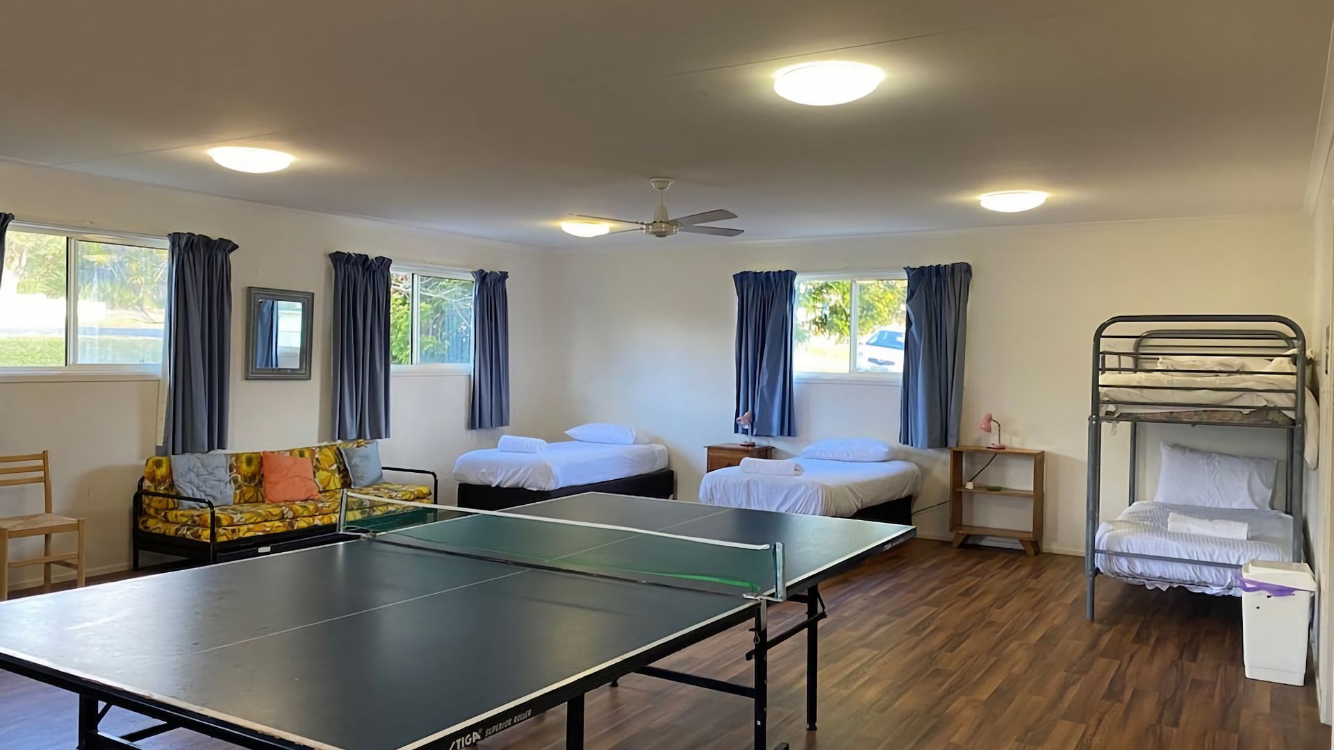 A room with a ping pong table, two beds, a bunk bed, a sofa, and a ceiling fan.