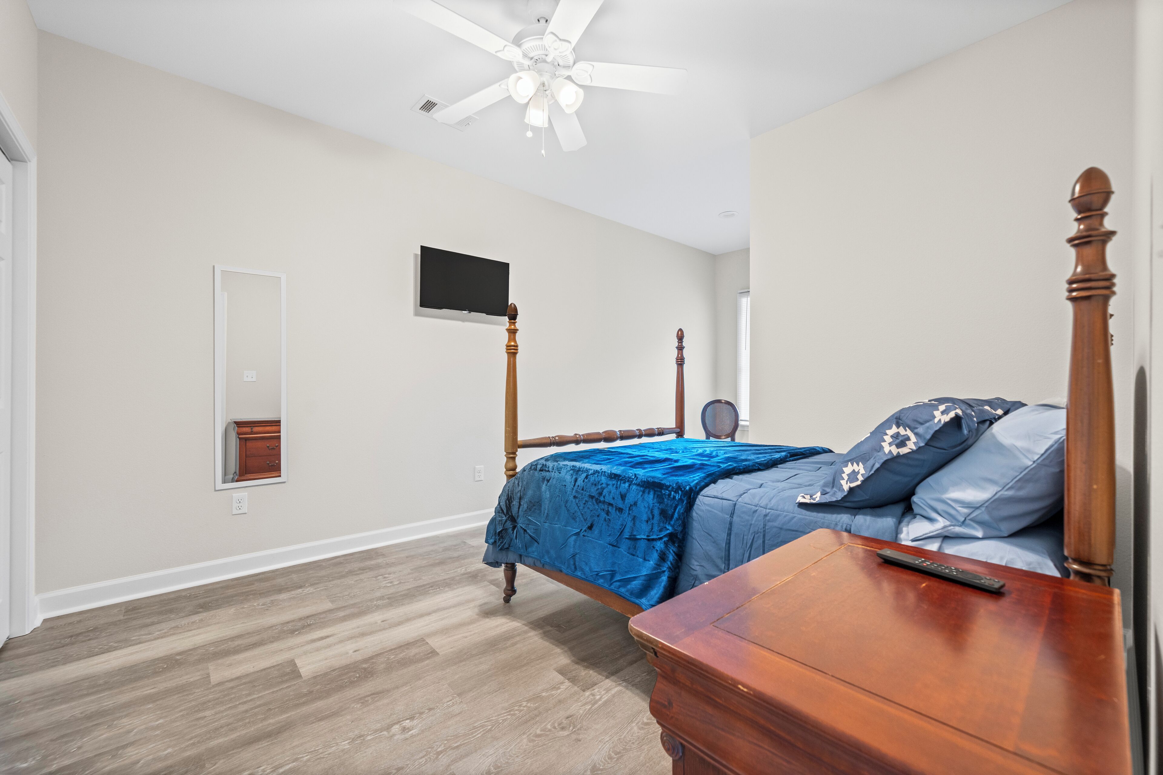 Une chambre avec un lit en bois, un ventilateur de plafond, un téléviseur fixé au mur et une commode en bois.