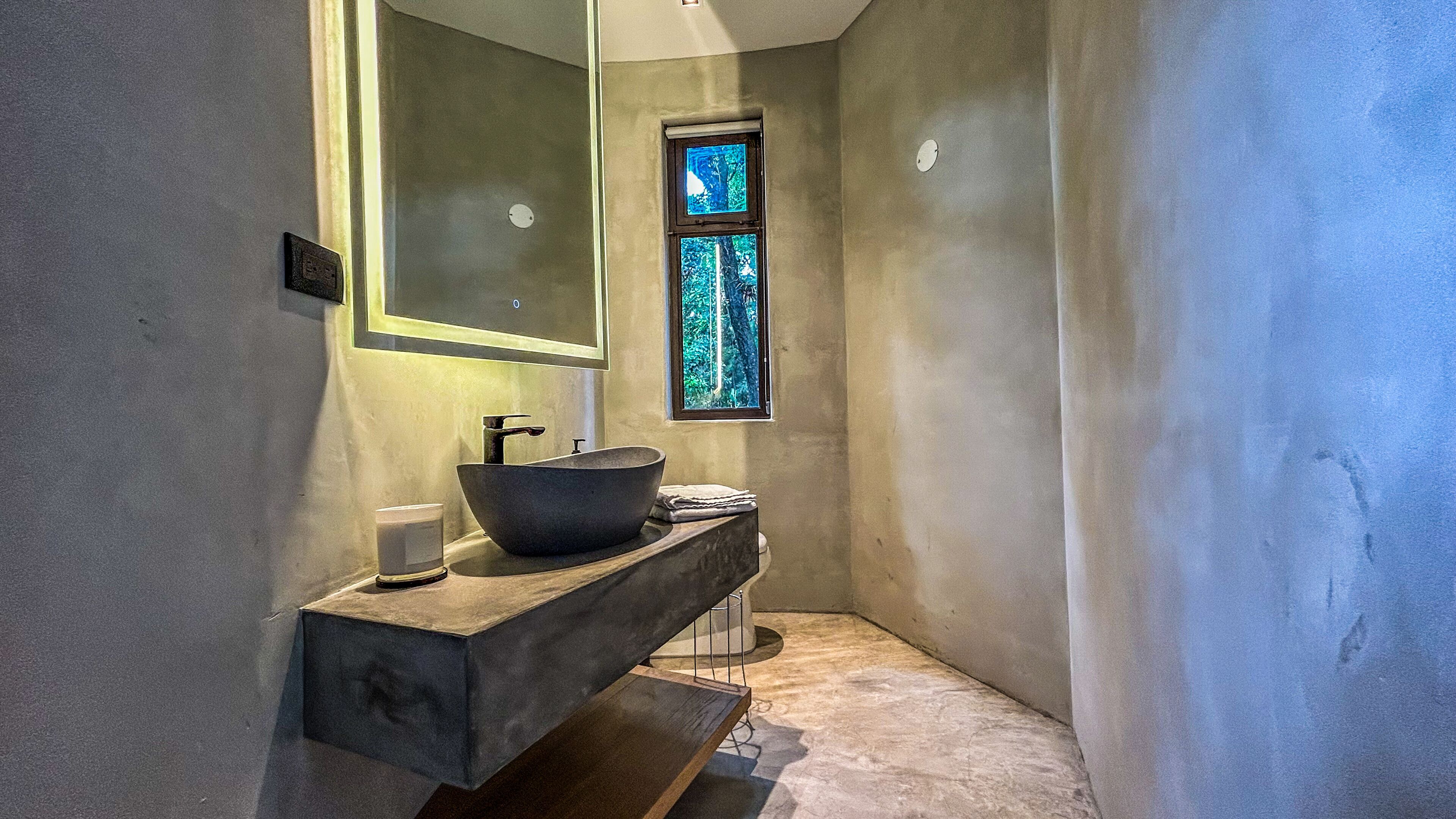 Un baño con un espejo grande, un lavabo independiente y un ventanal con vistas a árboles.