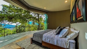 A bedroom with a large window overlooking a lush green landscape and a body of water.