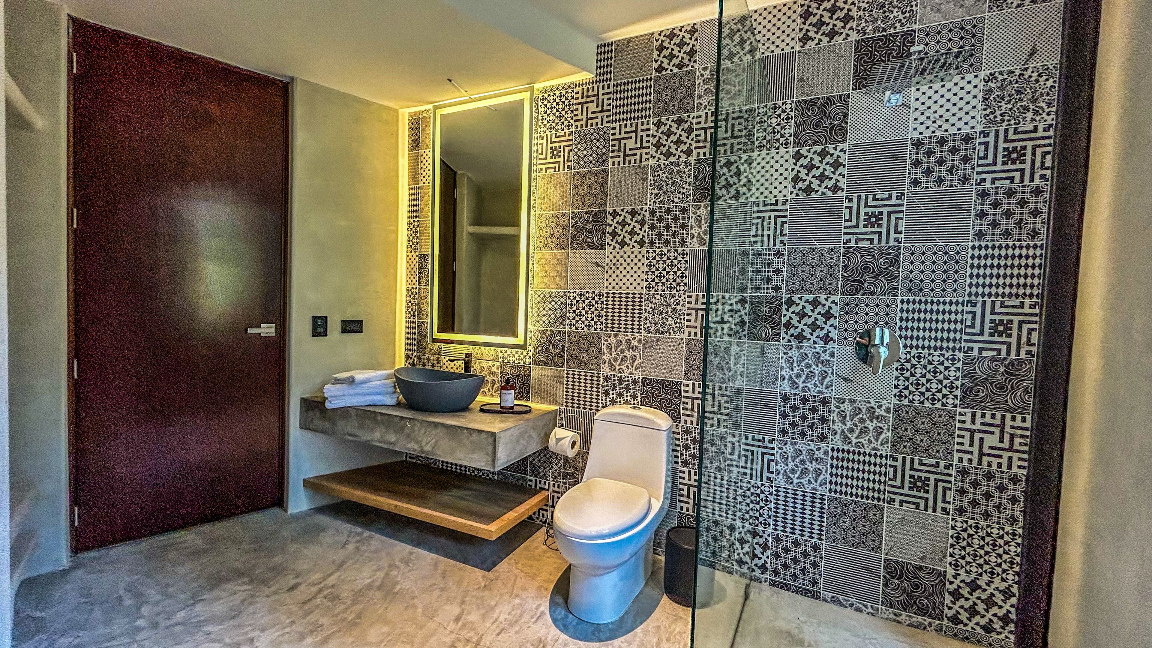 Une salle de bain au design moderne, dotée d’un grand miroir, d’un lavabo en forme de coupe et de toilettes. Les murs sont recouverts de carreaux à motifs.