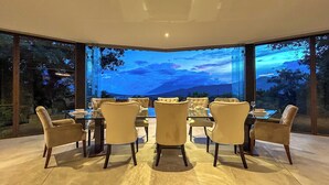 A dining area with a glass table and chairs, large windows with a view of mountains and trees, and a ceiling light.
