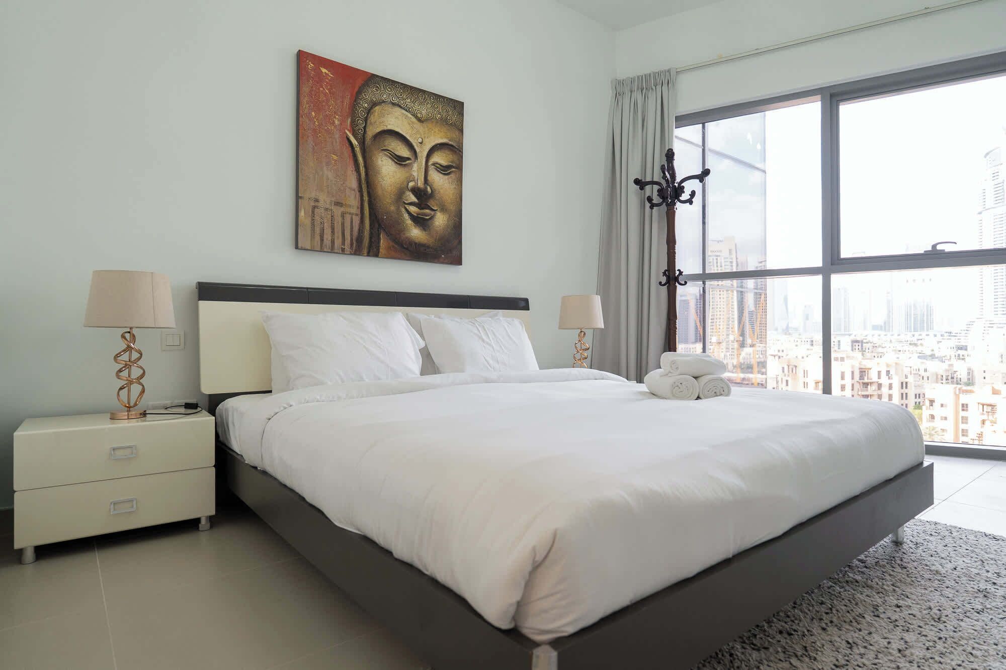 A modern bedroom with a large bed, two bedside tables with lamps, a city view through large windows, and a Buddha statue on the wall.