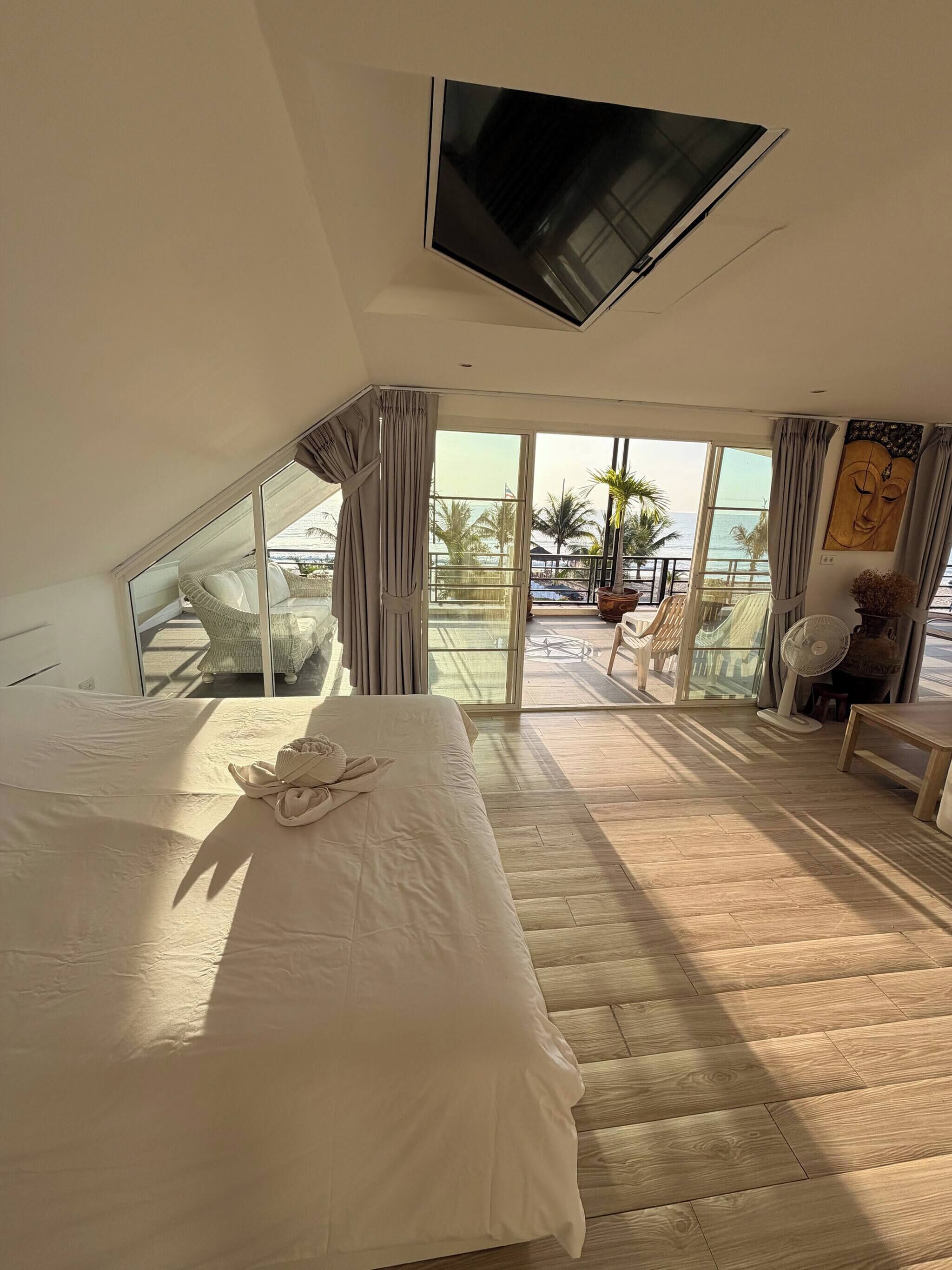 A bedroom with a large bed, a TV mounted on the ceiling, and a balcony with a view of the ocean.