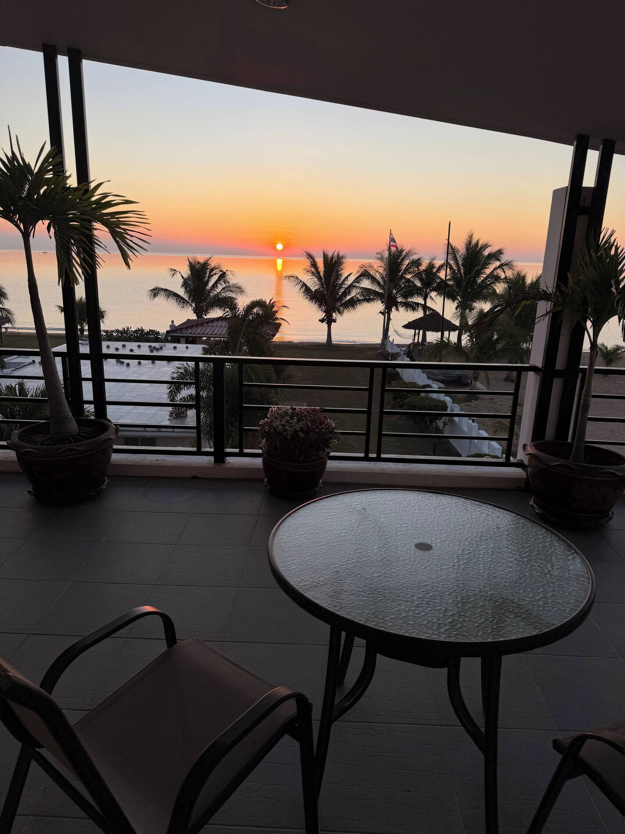 A balcony with a view of the sunset over the sea, potted plants, and a round table with chairs.