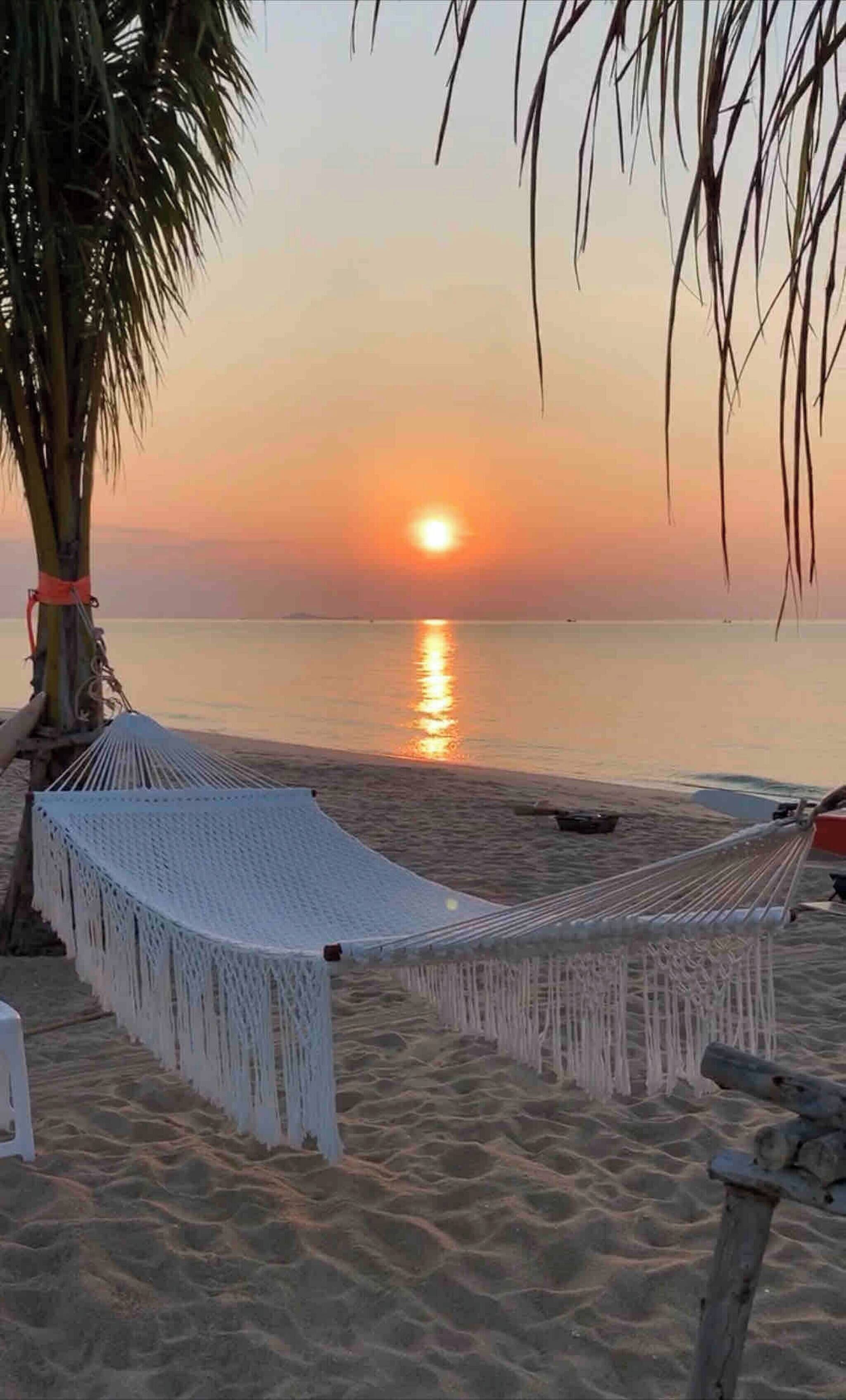 En hængekøje på en sandstrand med solnedgang i baggrunden.