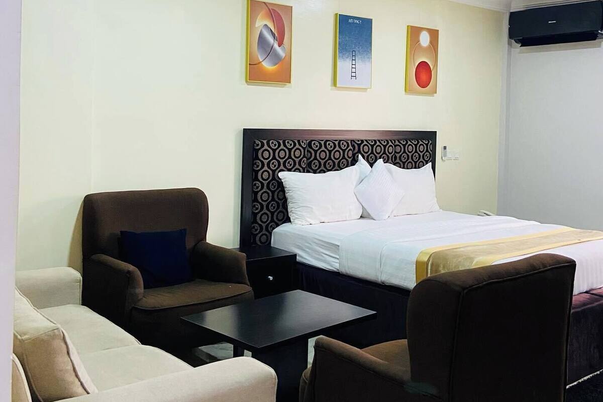 A hotel room with a bed, two armchairs, a small table, and a wall-mounted air conditioner.