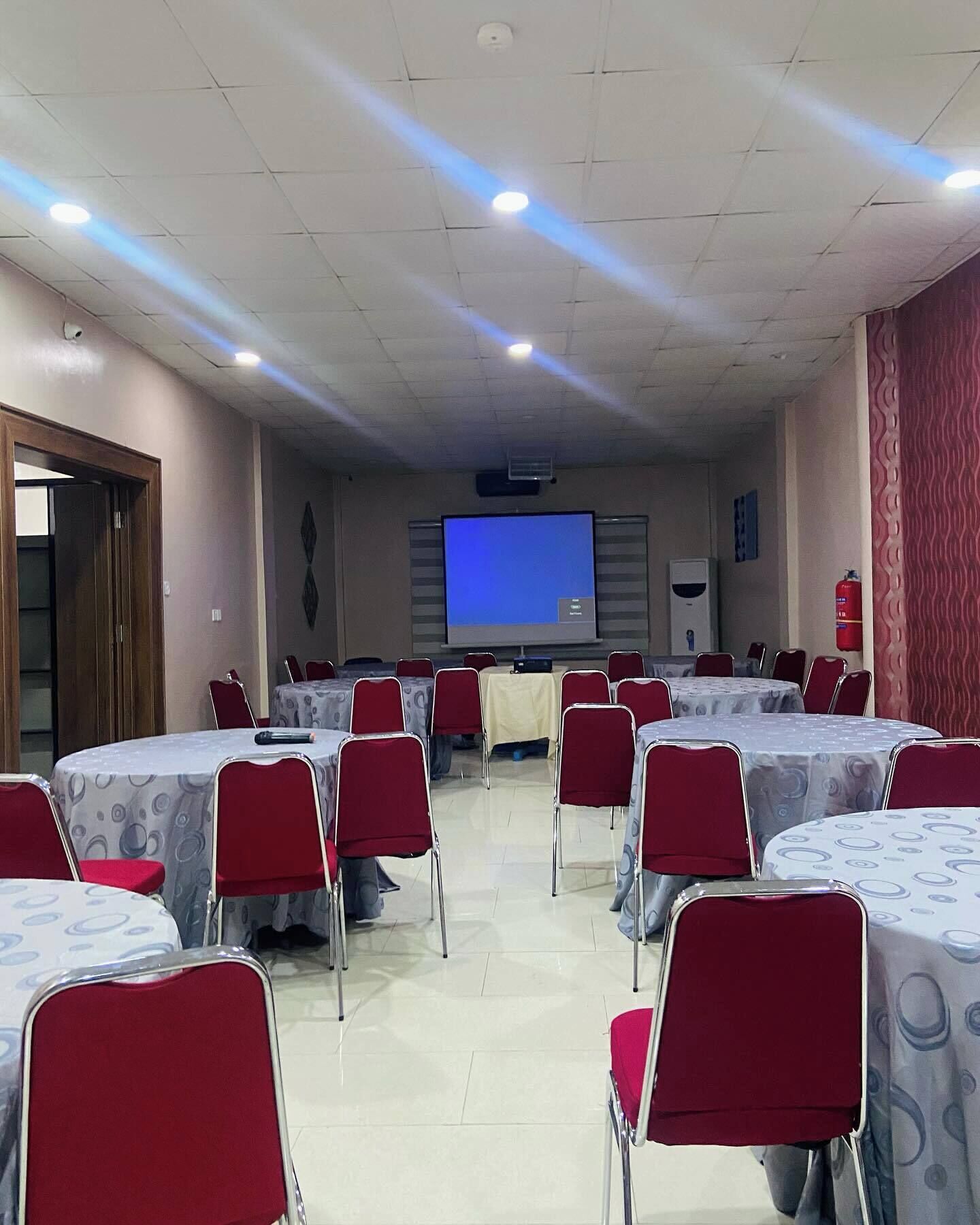 A conference room with multiple round tables and red chairs, a projector screen, and a fire extinguisher on the wall.