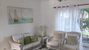 A living room with wicker furniture, a ceiling fan, a painting of sailboats, and a view of greenery outside. - Residencial El Carey! El lugar perfecto para tus vacaciones. (Punta Cana)