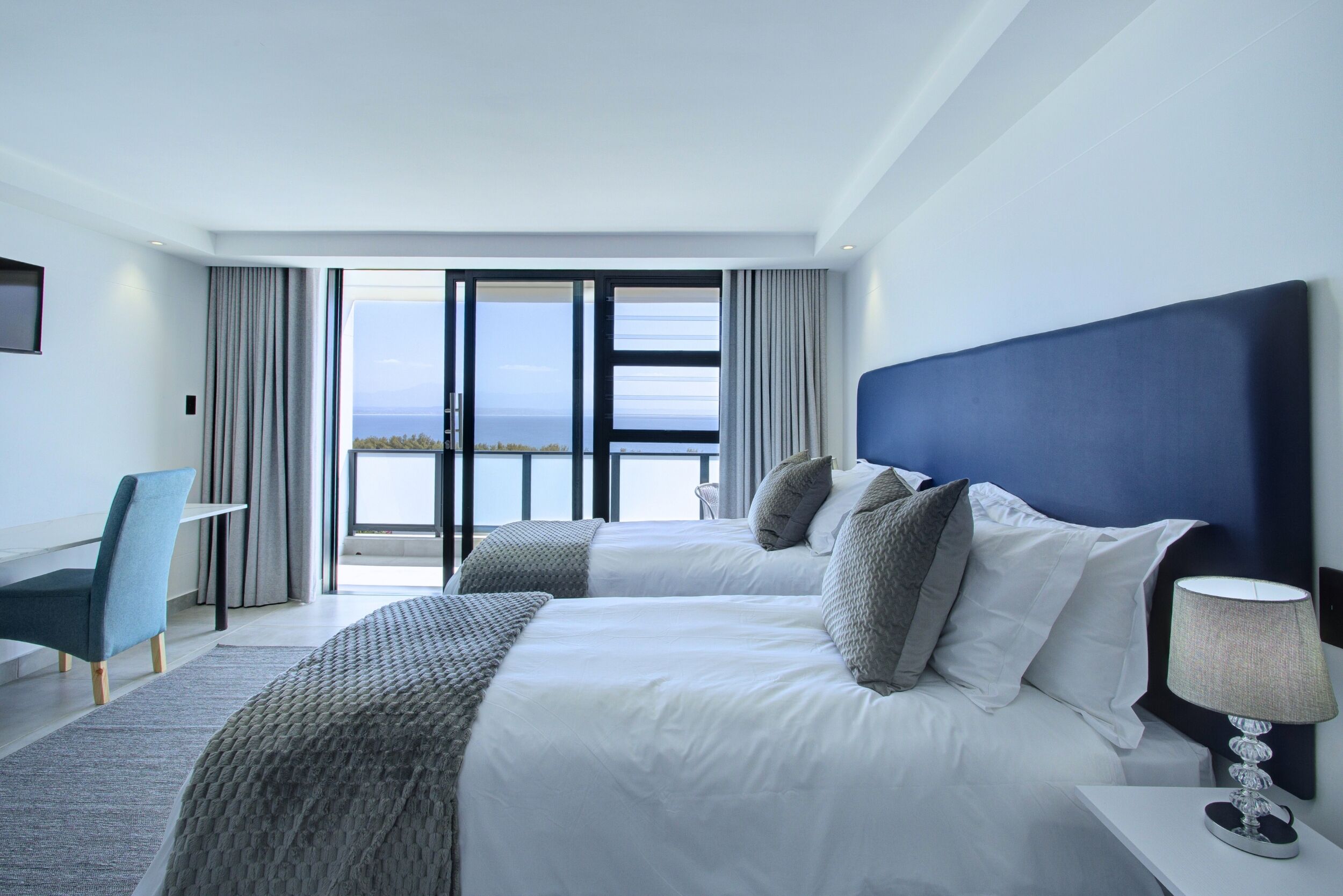 A modern hotel room with two beds, a desk, and a view of the sea.