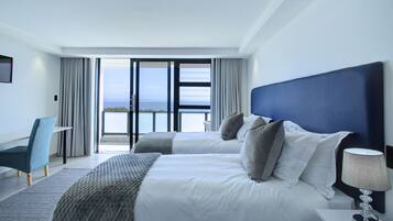 A modern hotel room with two beds, a desk, and a view of the sea.