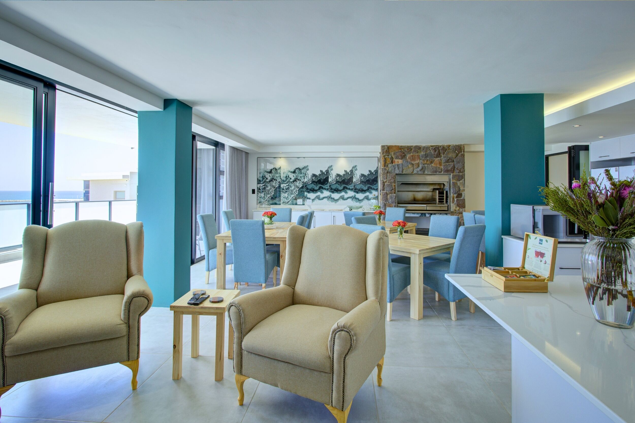 A modern living room with two beige armchairs, a dining table with blue chairs, a stone fireplace, and a large window overlooking a balcony.