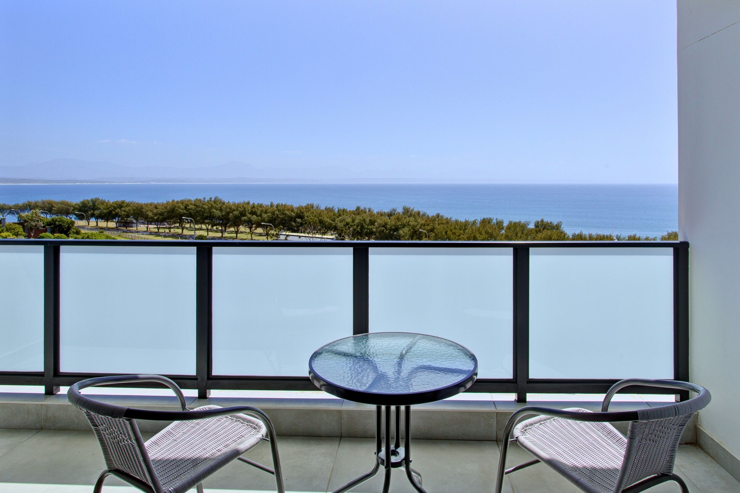 A balcony with a glass table and two wicker chairs, overlooking a coastal landscape with trees and the sea.