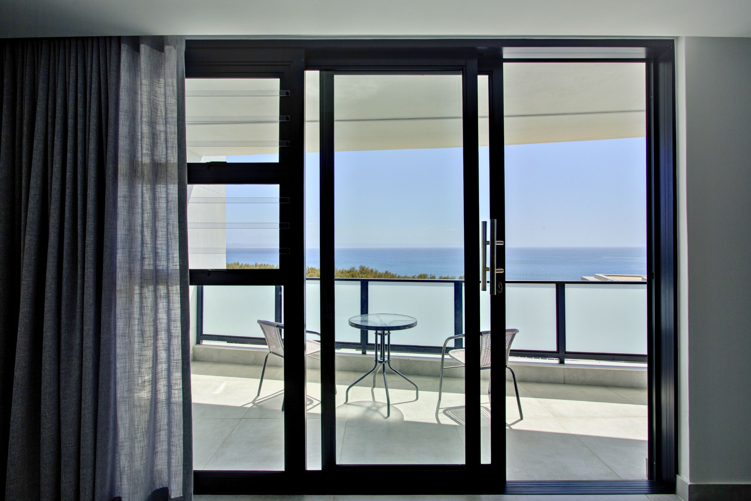 Un balcón con mesa y sillas, vista al mar y puerta corrediza de vidrio.