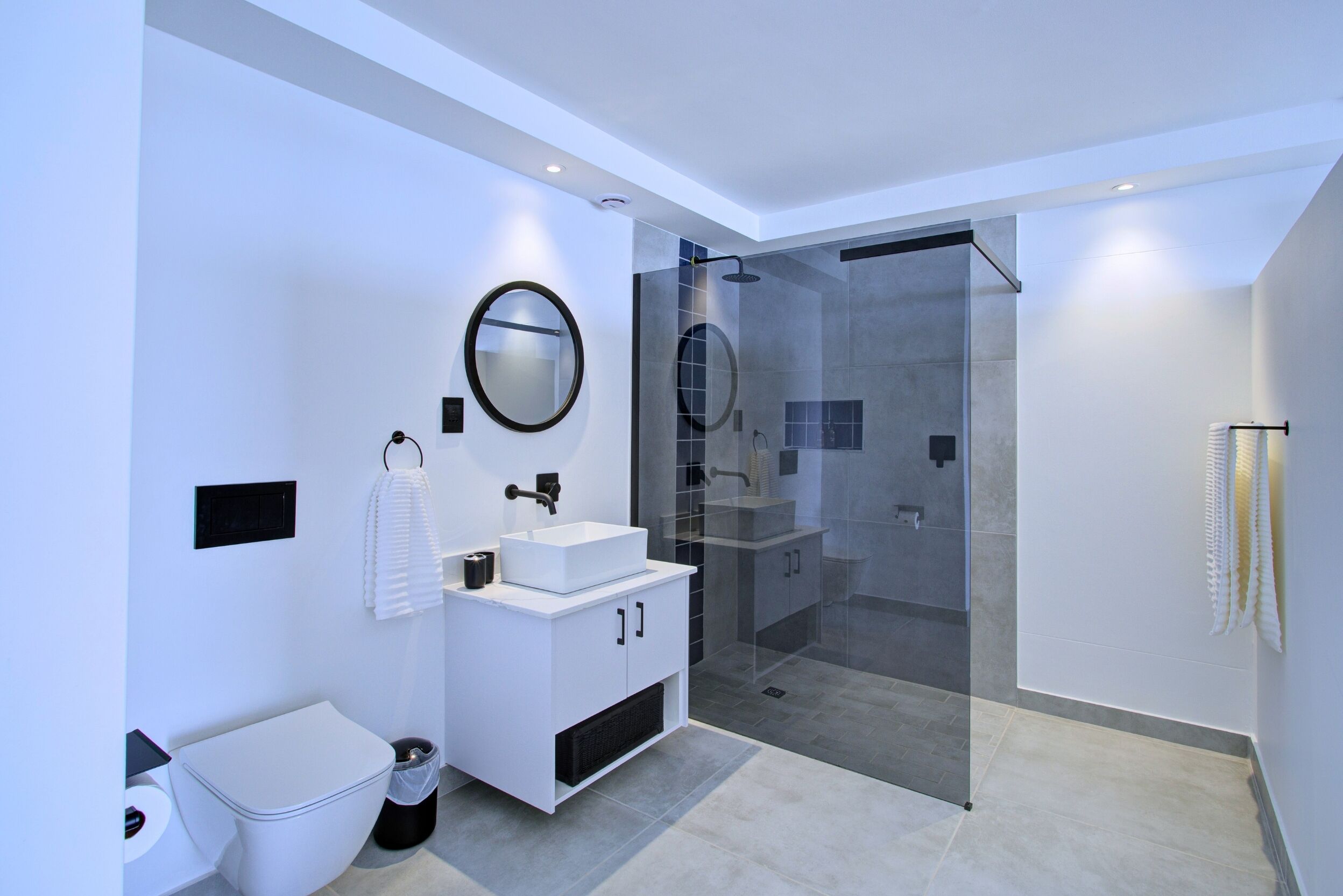 A modern bathroom with a white vanity, a round mirror, a glass-enclosed shower, and a toilet.
