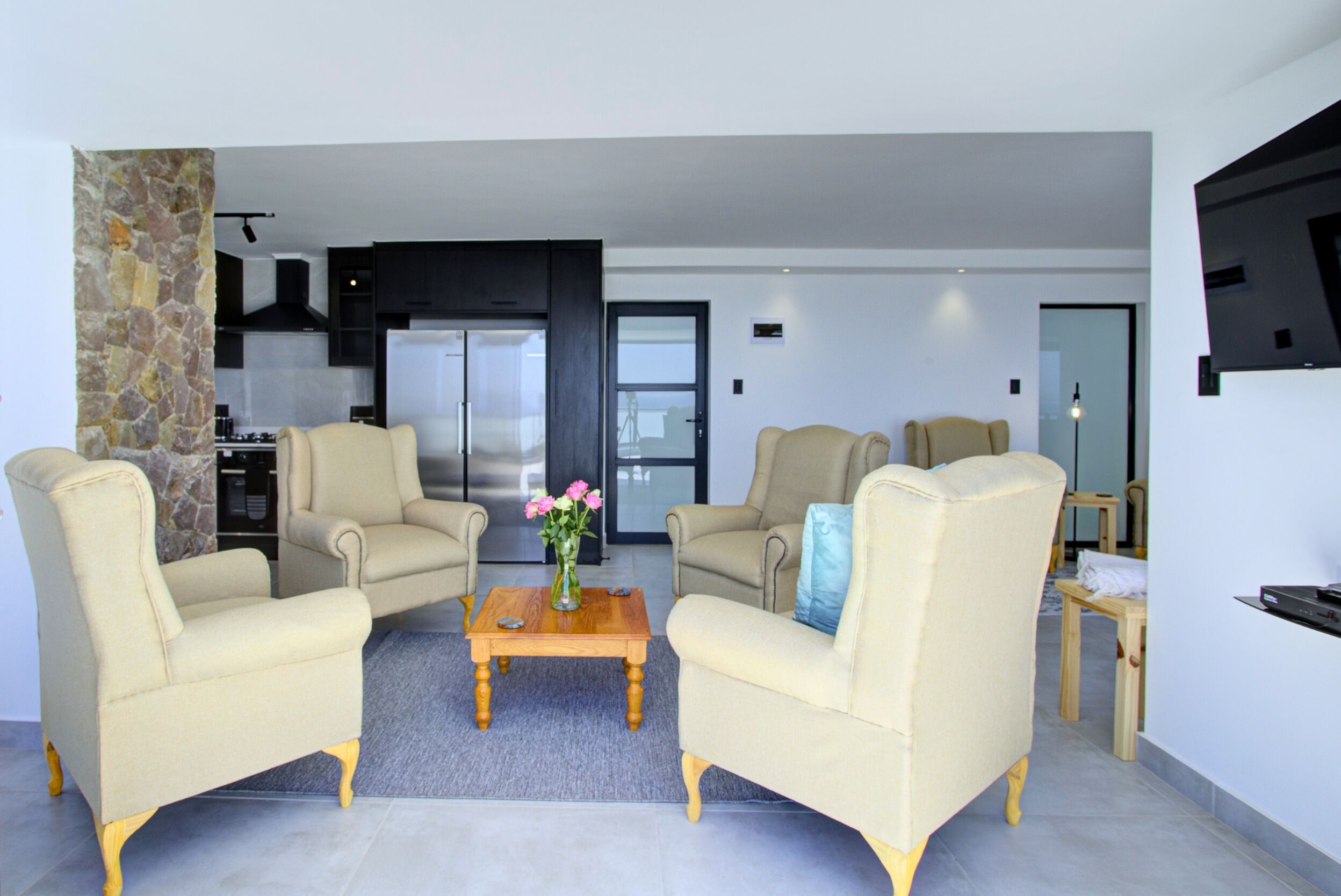 A modern living room with two beige armchairs, a wooden coffee table with a vase of flowers, and a kitchen in the background.