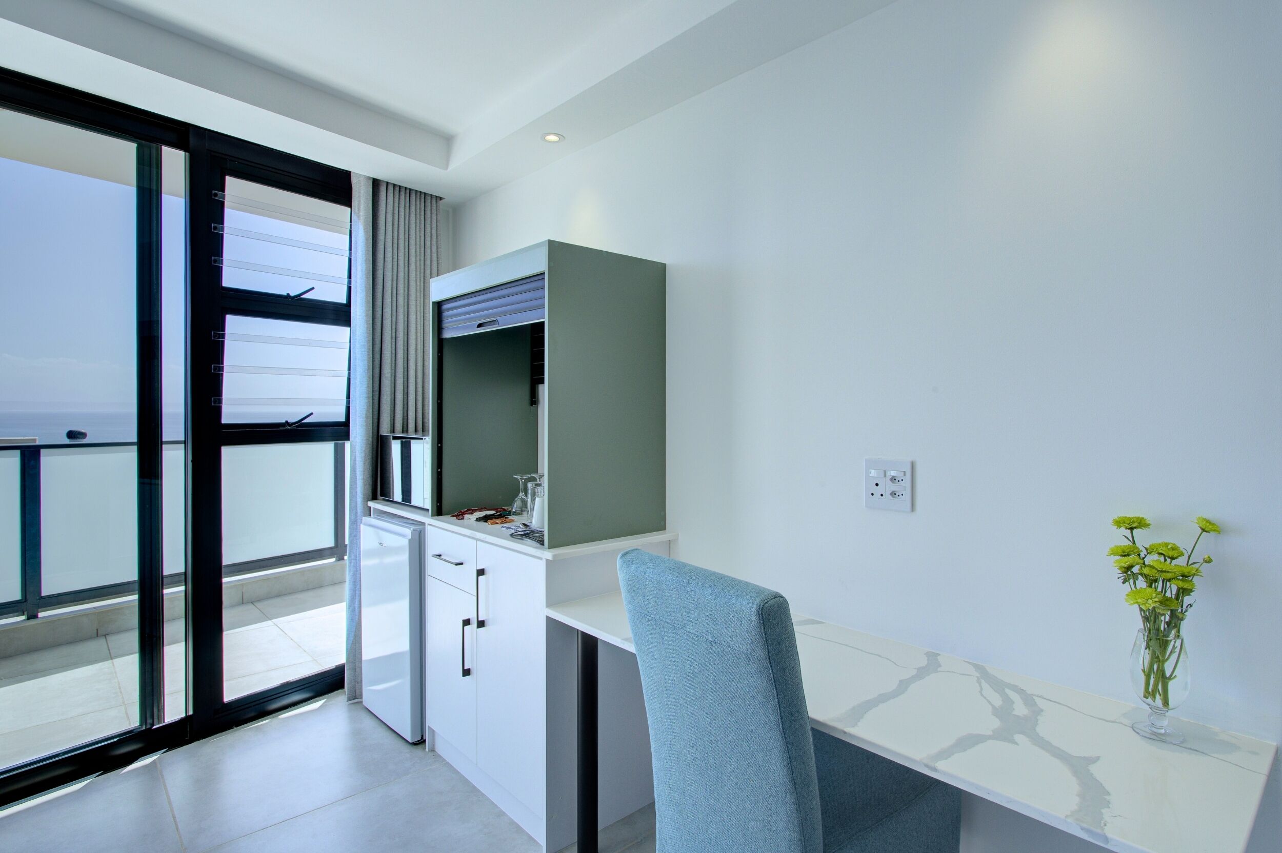 Ein modernes Zimmer mit weißem Schreibtisch, blauem Sessel, einer kleinen Küchenzeile und einem großen Fenster.