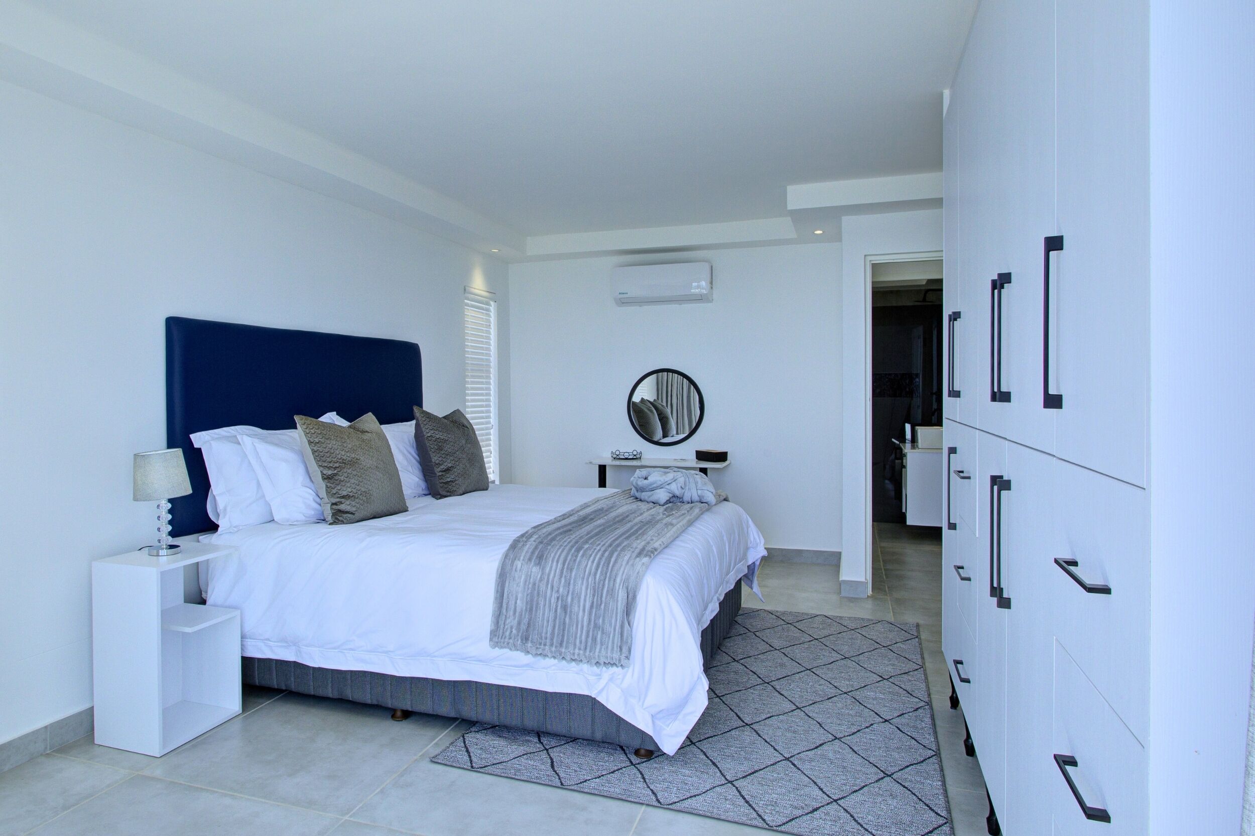 A modern bedroom with a large bed, white nightstands, a mirror, and a wardrobe with multiple drawers.