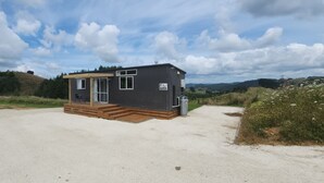 A small, dark-colored cabin with a wooden deck and a single window, surrounded by a gravel path and grassy areas.