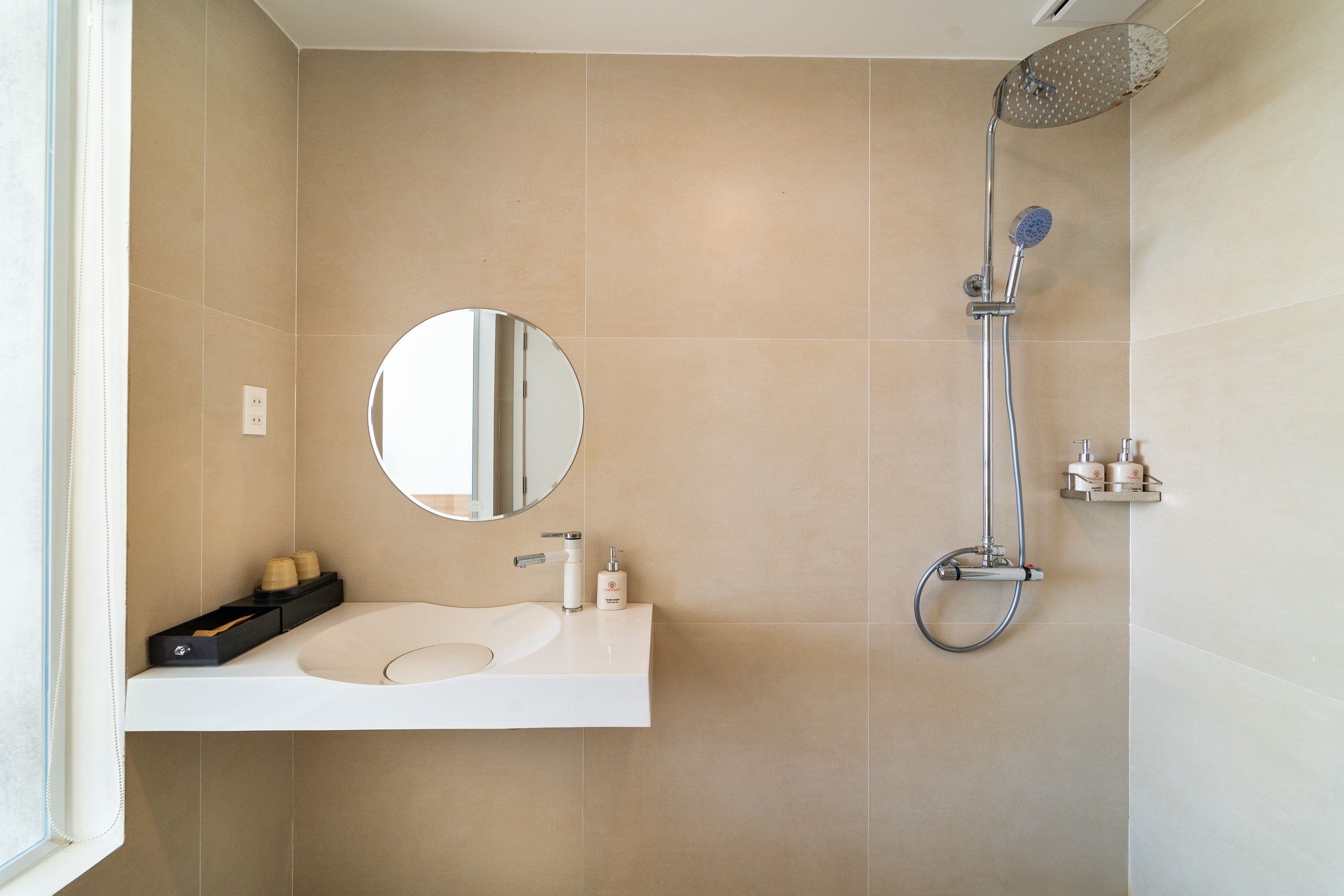 Un baño moderno con un lavamanos blanco, un espejo redondo, un cabezal de ducha manual y un inodoro de pared.
