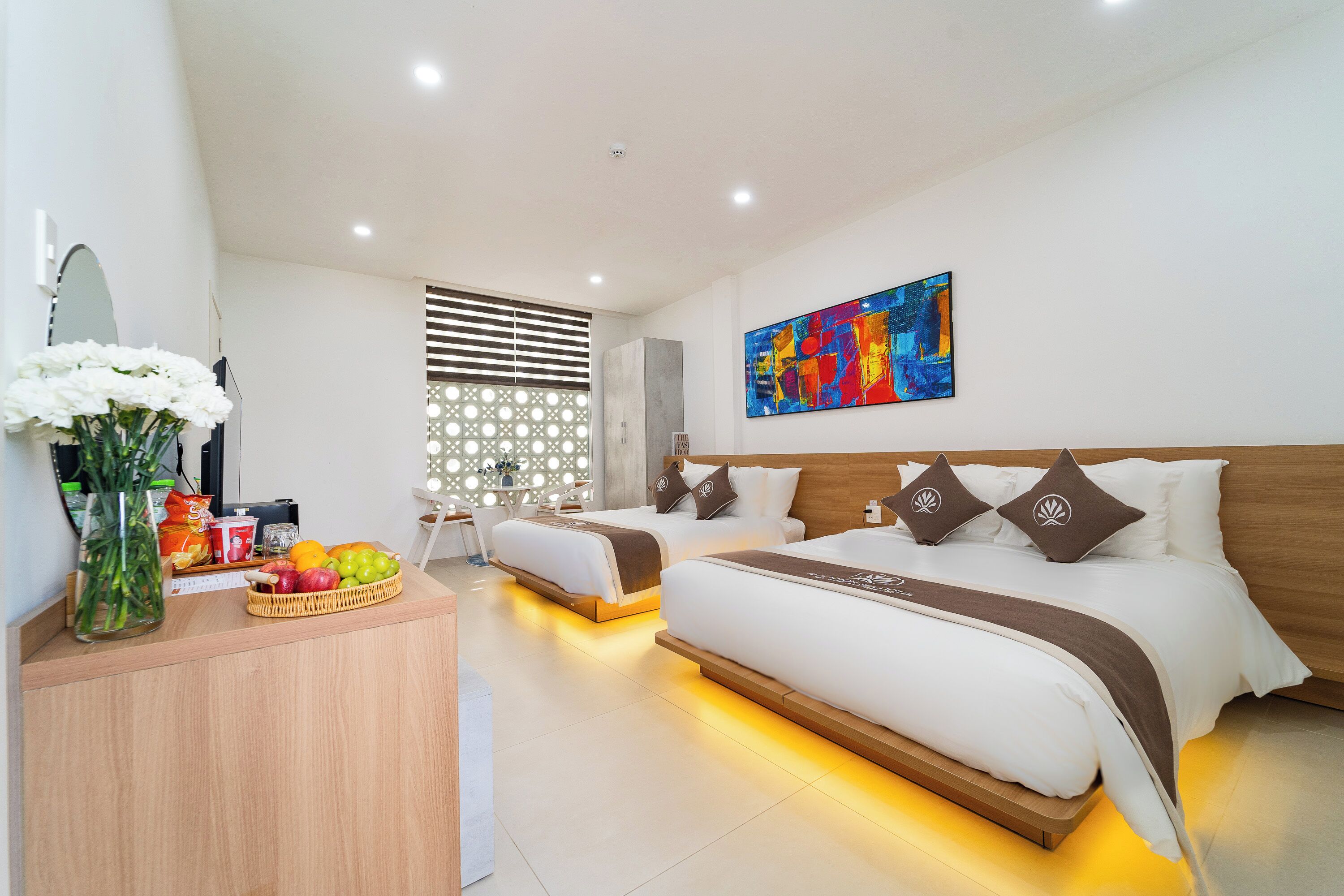A modern hotel room with a large bed, a desk with a fruit basket, and a colorful abstract painting on the wall.