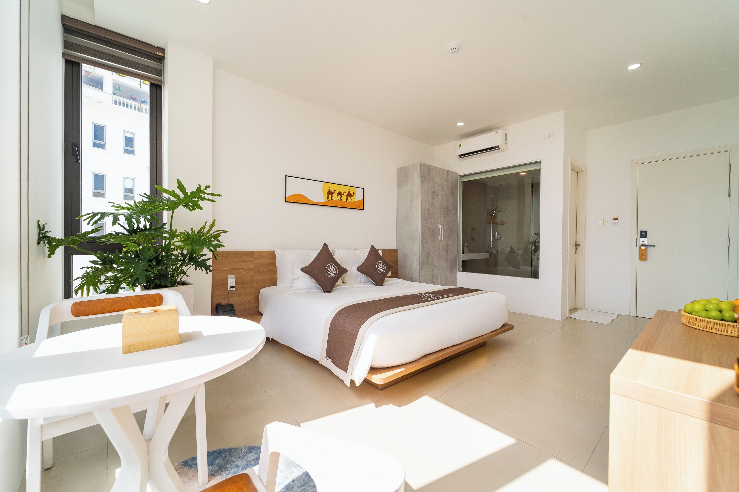 A modern hotel room with a bed, a dining table, a plant, and a view of the city.
