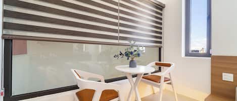 Una habitación moderna con un ventanal grande cubierto por persianas horizontales, una pequeña mesa redonda con dos sillas, y un florero con flores sobre la mesa.