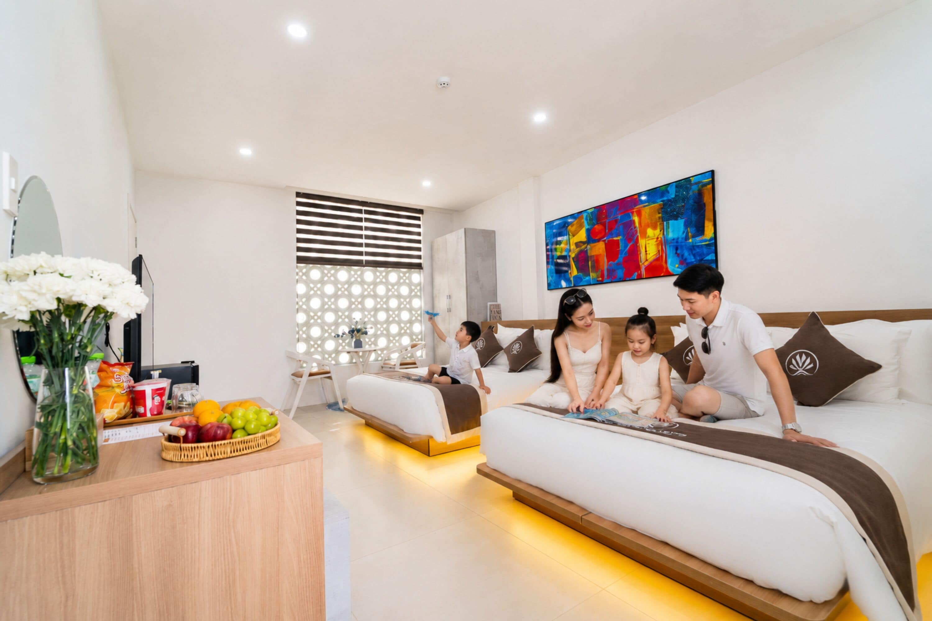 Camera d'hotel moderna con un letto grande, un divano più piccolo, un tavolo da pranzo con frutta e un quadro astratto dai colori vivaci.
