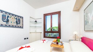 A hotel room with a bed, a bedside table with a tray of breakfast, a window with a view of greenery, a decorative wall hanging, and a bedside lamp. - Il Vecchio Faro 66 (Loiri Porto San Paolo)