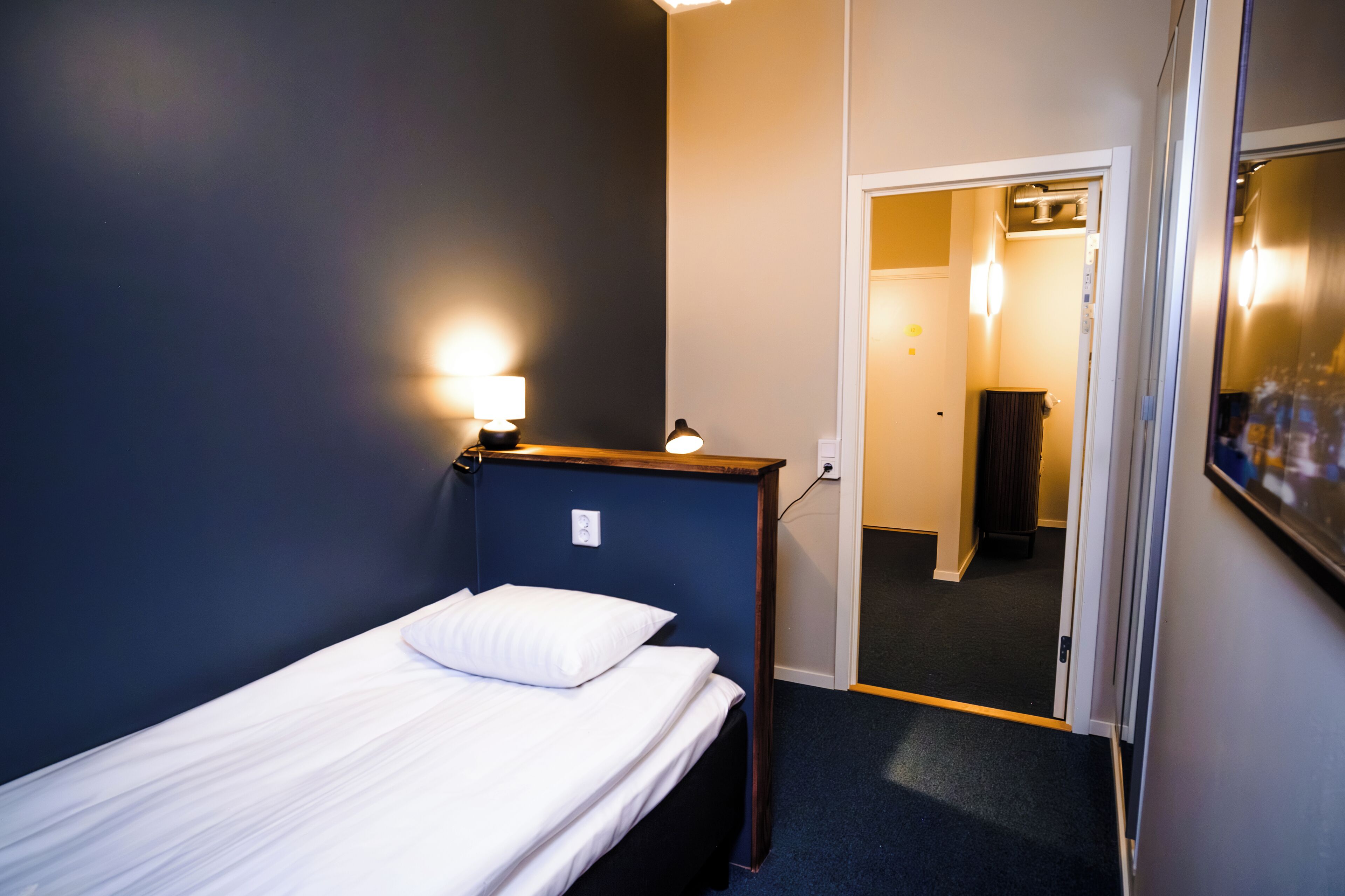A small, neatly made bed with a wooden headboard and a white pillow in a room with dark blue walls and a carpeted floor.