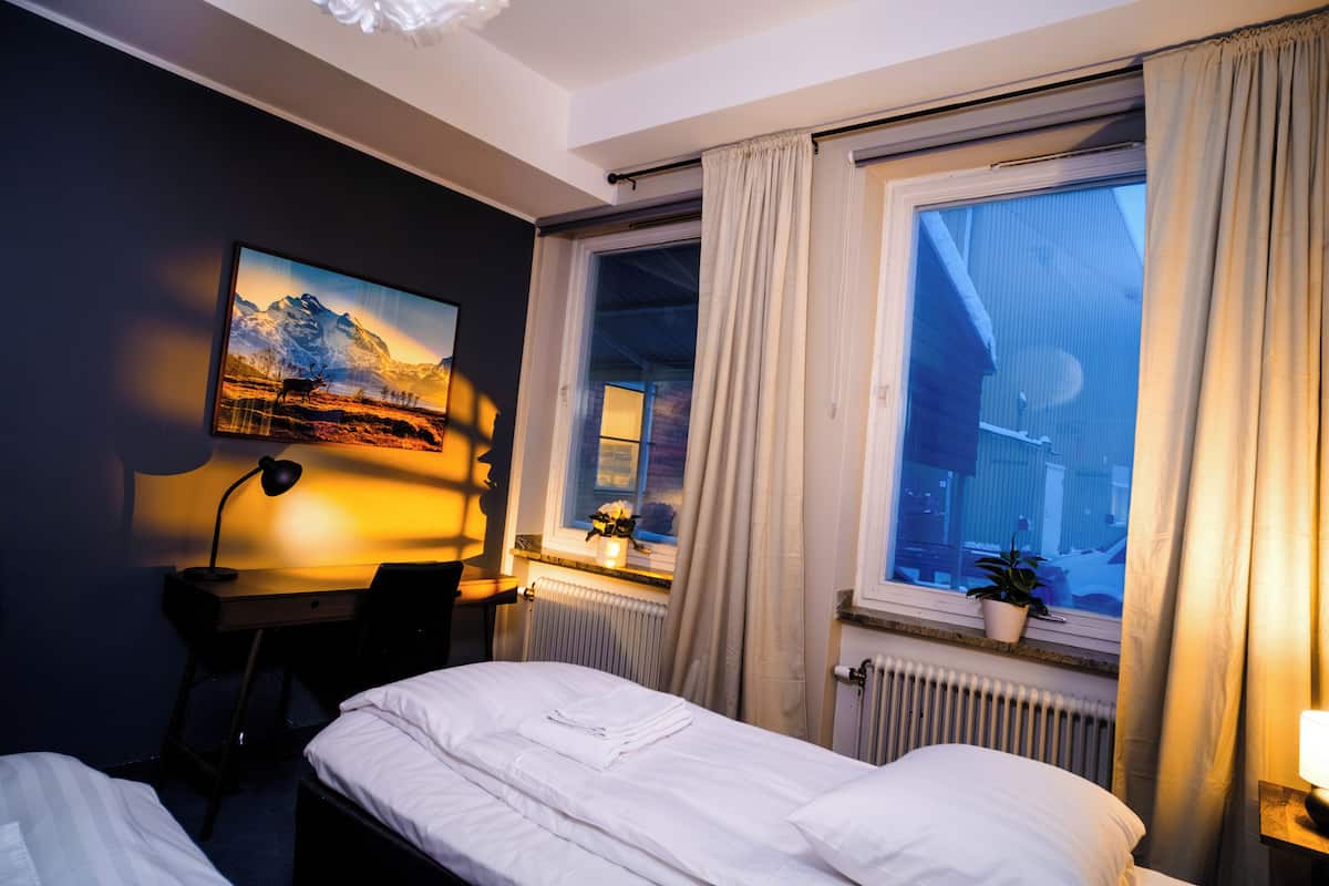 Une chambre d’hôtel avec un lit, un bureau, une chaise, une fenêtre avec des rideaux, un radiateur et un tableau au mur.
