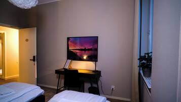 A bedroom with two beds, a desk with a computer, a painting on the wall, and a window with a view.