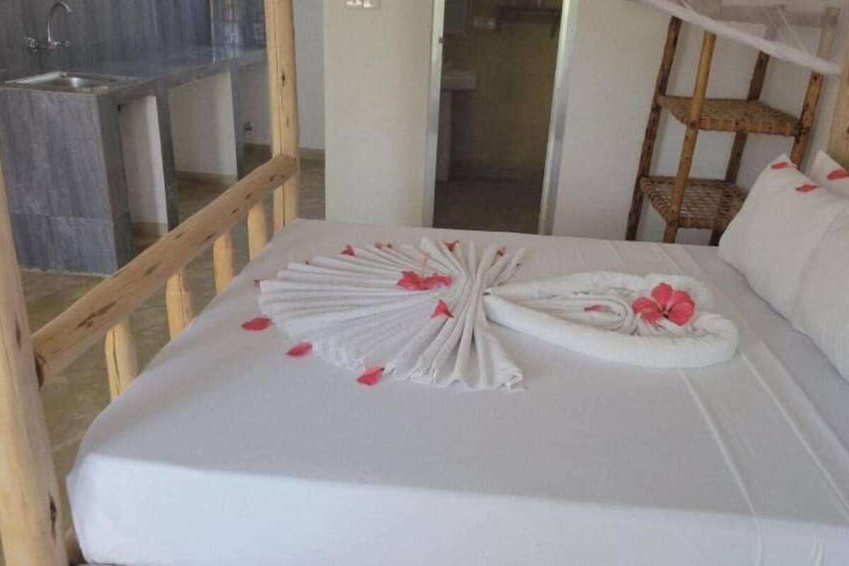 Ein ordentlich bezogenes Bett mit weißen Leinentüchern und rosa Blütenblättern, ein Holzstuhl und eine kleine Holzablage.