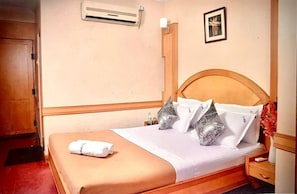A hotel room with a bed, a wooden headboard, a nightstand, a bedside table, a framed picture on the wall, and an air conditioning unit. - HOTEL TAJ MAHAL (Bengaluru)