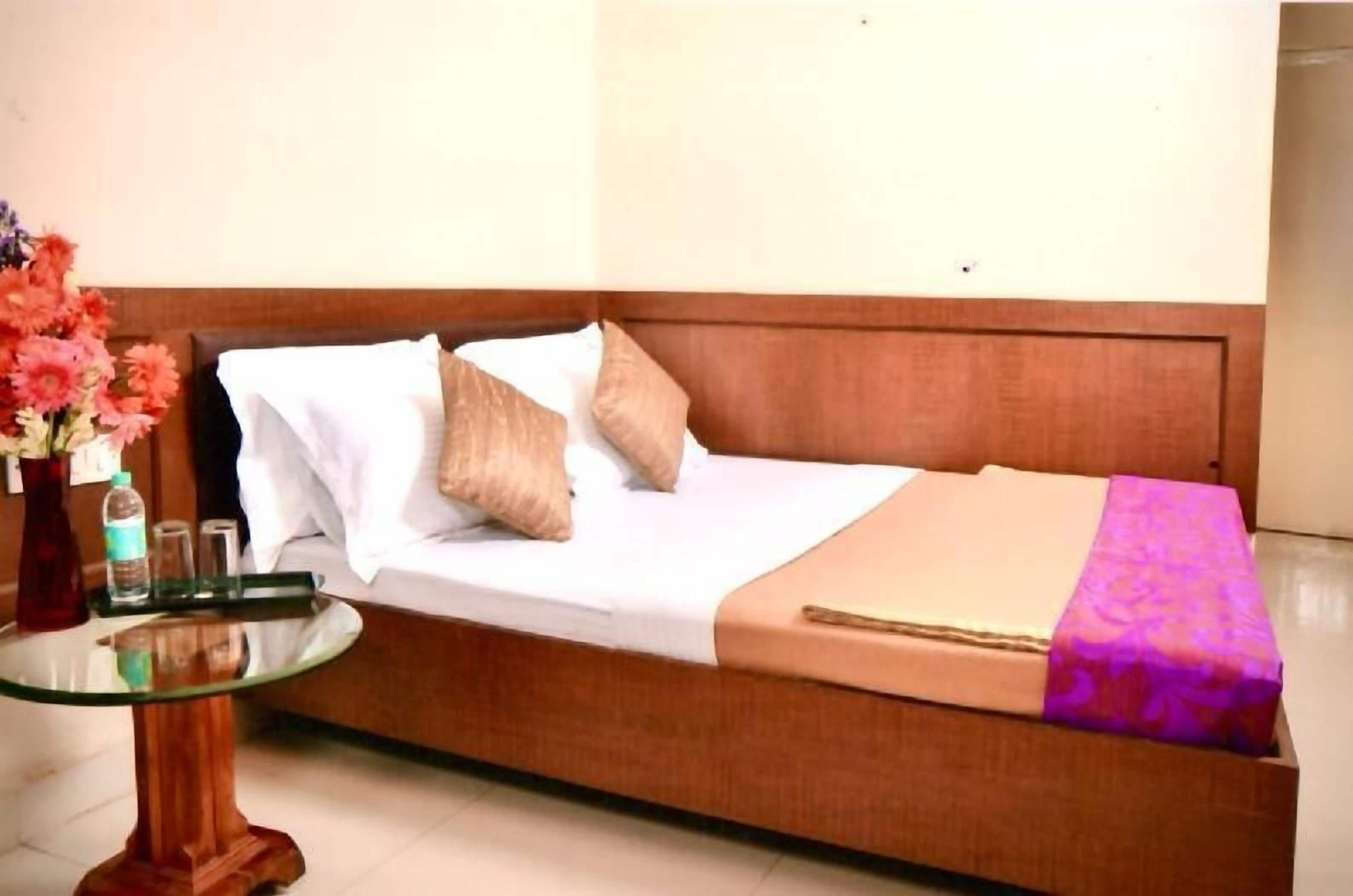 A single bed with a wooden headboard, a glass table with a vase of flowers, and a small table with bottles and glasses.