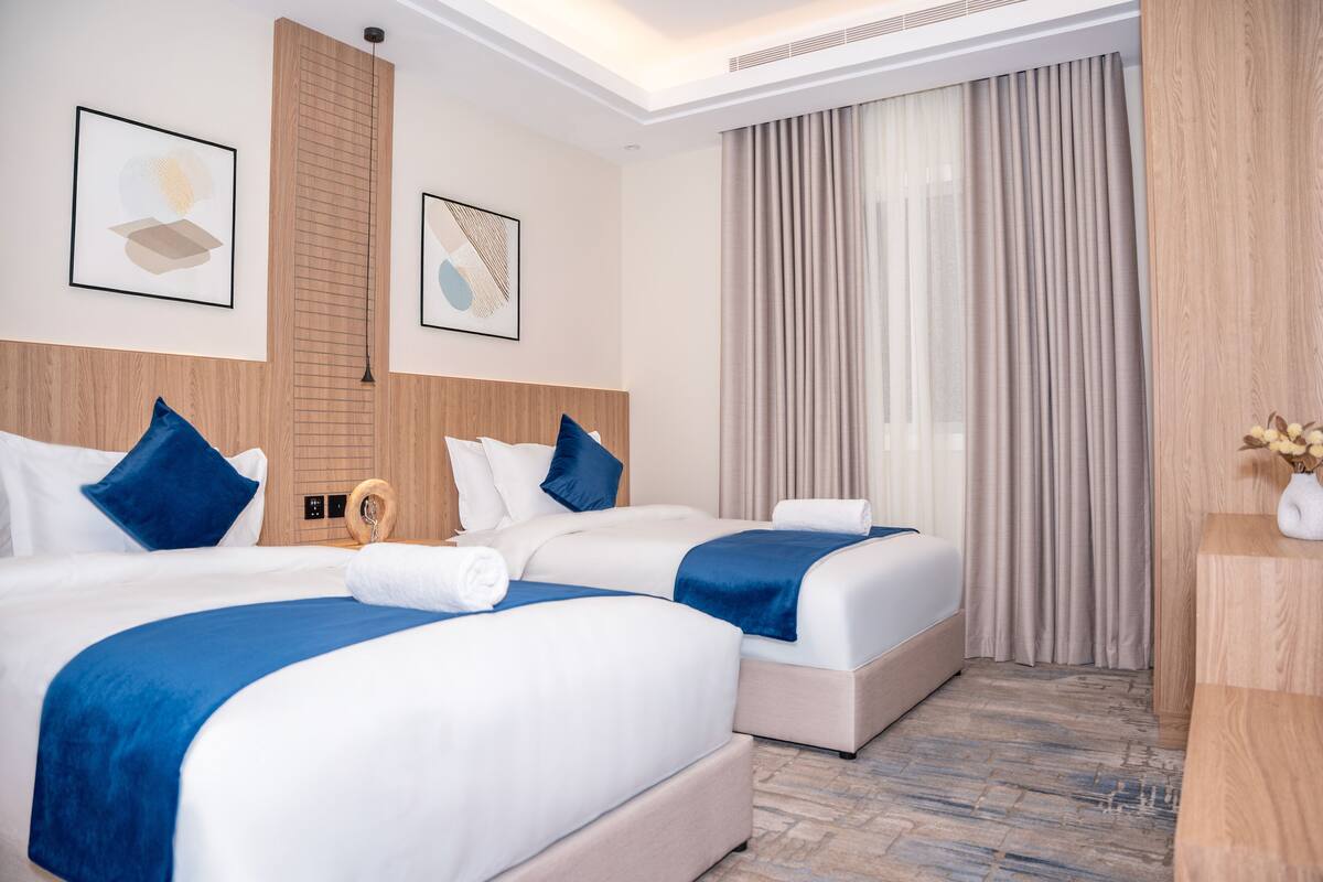 A hotel room with two beds, a wooden headboard, a large window with curtains, and artwork on the walls.