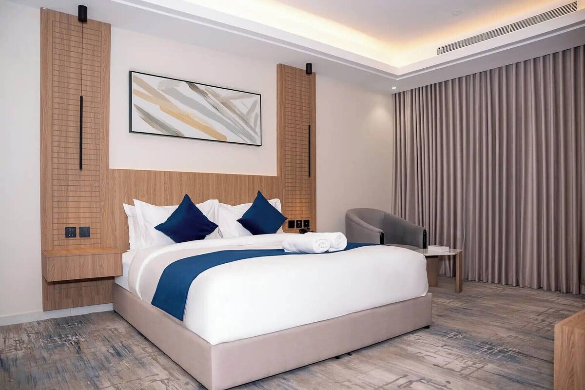 A modern hotel room with a large bed, a headboard with a built-in shelf, a bedside table, a chair, and a large window with curtains.