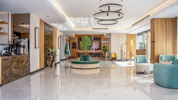 A modern hotel lobby with a coffee station, a reception desk, a green seating area, and a large window.