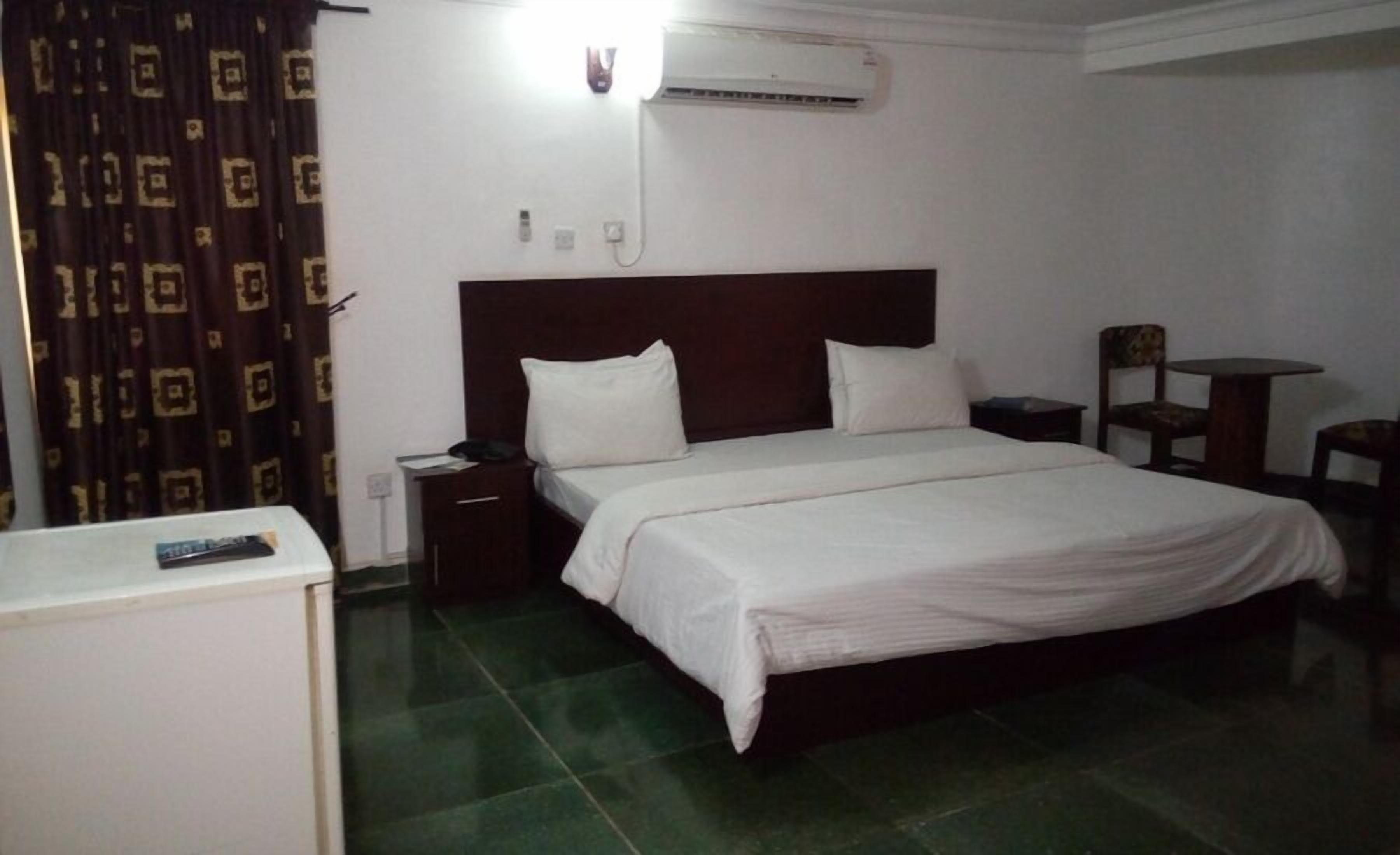 A hotel room with a bed, bedside tables, an air conditioning unit, a small table with chairs, and a curtain with a geometric pattern.