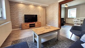 Un salon avec un mur en pierre, un meuble de rangement en bois pour l’équipement audiovisuel, un téléviseur à écran plat, une table basse en bois et un canapé gris.