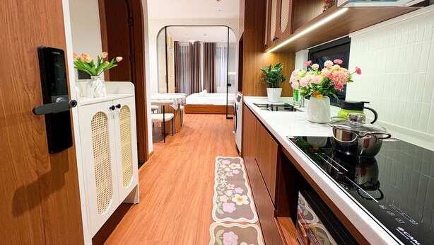 A compact kitchen with a stove, sink, and cabinets, leading to a bedroom with a bed and curtains. - GAIA House Le Duan (Hanoi)