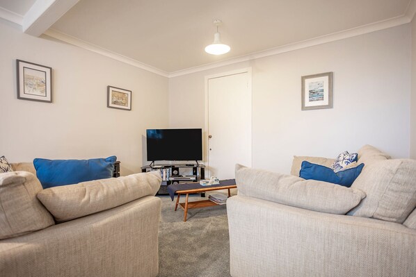 A living room with two beige sofas, a coffee table, a flat-screen TV, and framed pictures on the walls. - Seascape Cottage at Crail Harbour (Crail)