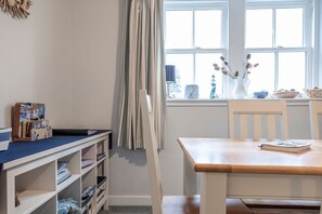 A room with a wooden table, white shelves, a window with curtains, and a vase with dried flowers. - Seascape Cottage at Crail Harbour (Crail)