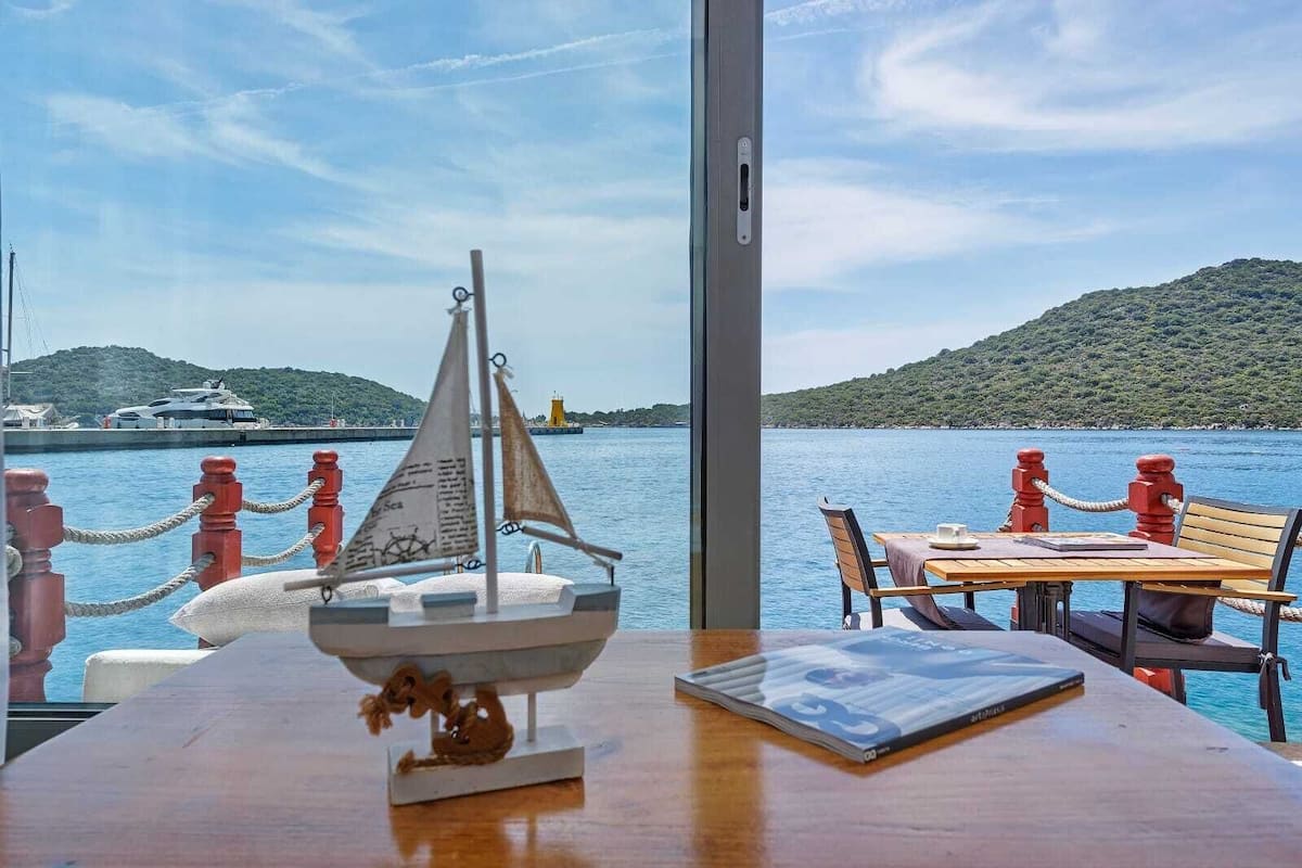 A table with a model sailboat, a book, and a cup, overlooking a harbor with boats and a hilly landscape.